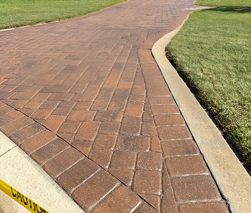 A winding brick pathway bordered by neatly trimmed grass, with a caution tape warning in the foreground.